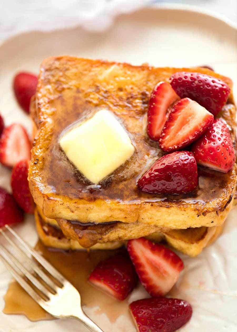 Pile of French Toast on a rustic white plate, doused with maple syrup and topped with a pat of melting butter and strawberries.