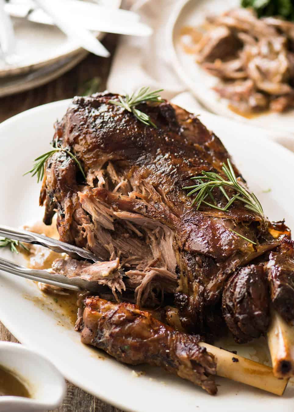 A Slow Roasted Lamb Leg on a white platter with some of the tender meat pulled off with tongs. Garnished with sprigs of rosemary.