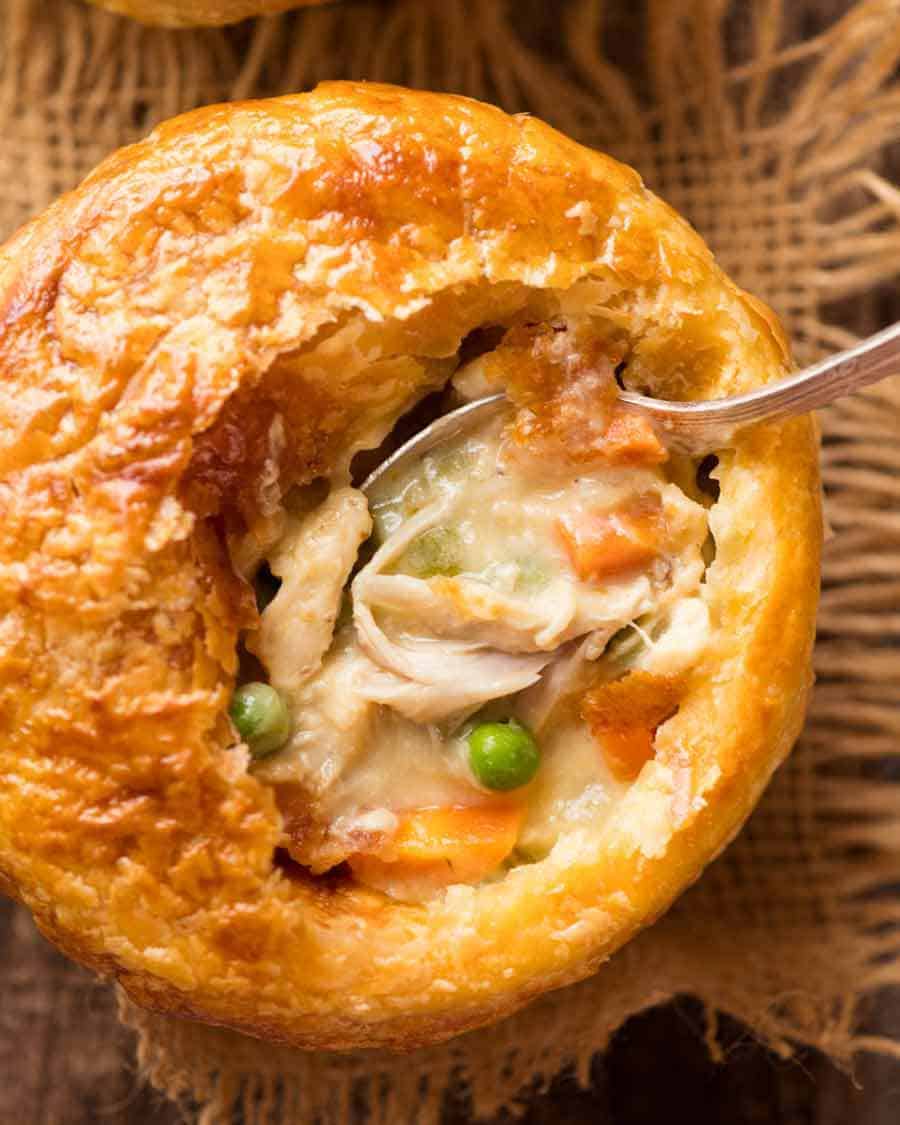Close up of spoon shopping up filling of Chicken Pot Pie in a ramekin with puff pastry topping, made from scratch with uncooked chicken.